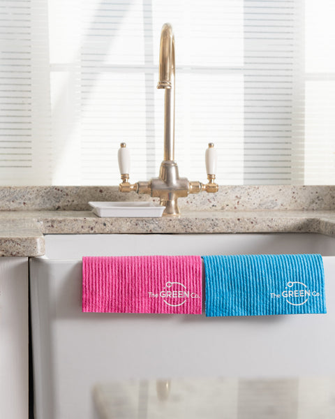 Pink cellulose dishcloth and blue cellulose dishcloth draped over ceramic sink with golden taps