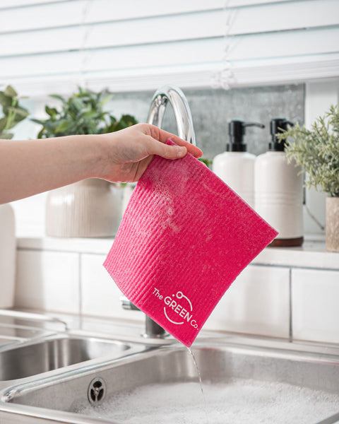 Pink Swedish Dish Cloth dripping of soapy water, being held over filled sink with bubbled in a kitchen environment