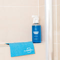 Blue Bathroom Cleaner GreenTab in Forever Bottle and Blue Cellulose Dish Cloth sitting on the edge of a Shower