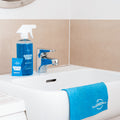 Blue Bathroom Cleaner GreenTab in Forever Bottle and Blue GreenTab in Packaging sitting next to a tap on Bathroom Sink and Blue Cellulose Dish Cloth hanging over the Bathroom Sink