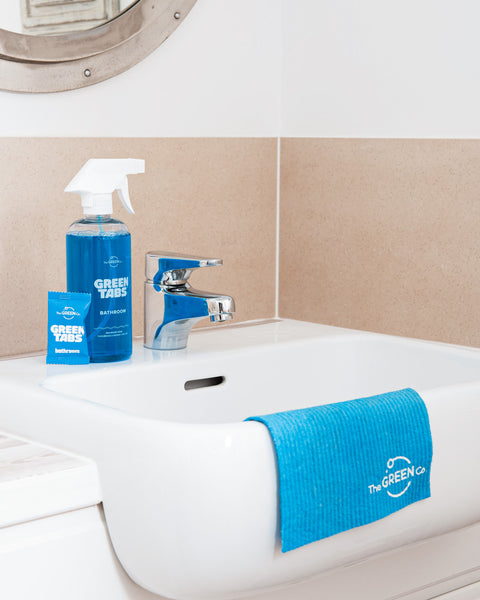 Blue Bathroom Cleaner GreenTab in Forever Bottle and Blue GreenTab in Packaging sitting next to a tap on Bathroom Sink and Blue Cellulose Dish Cloth hanging over the Bathroom Sink