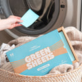 Blue Fresh Linen Fabric conditioner GreenSheets getting put into the washing machine by a hand. Packaging lying on top of washing basket with laundry